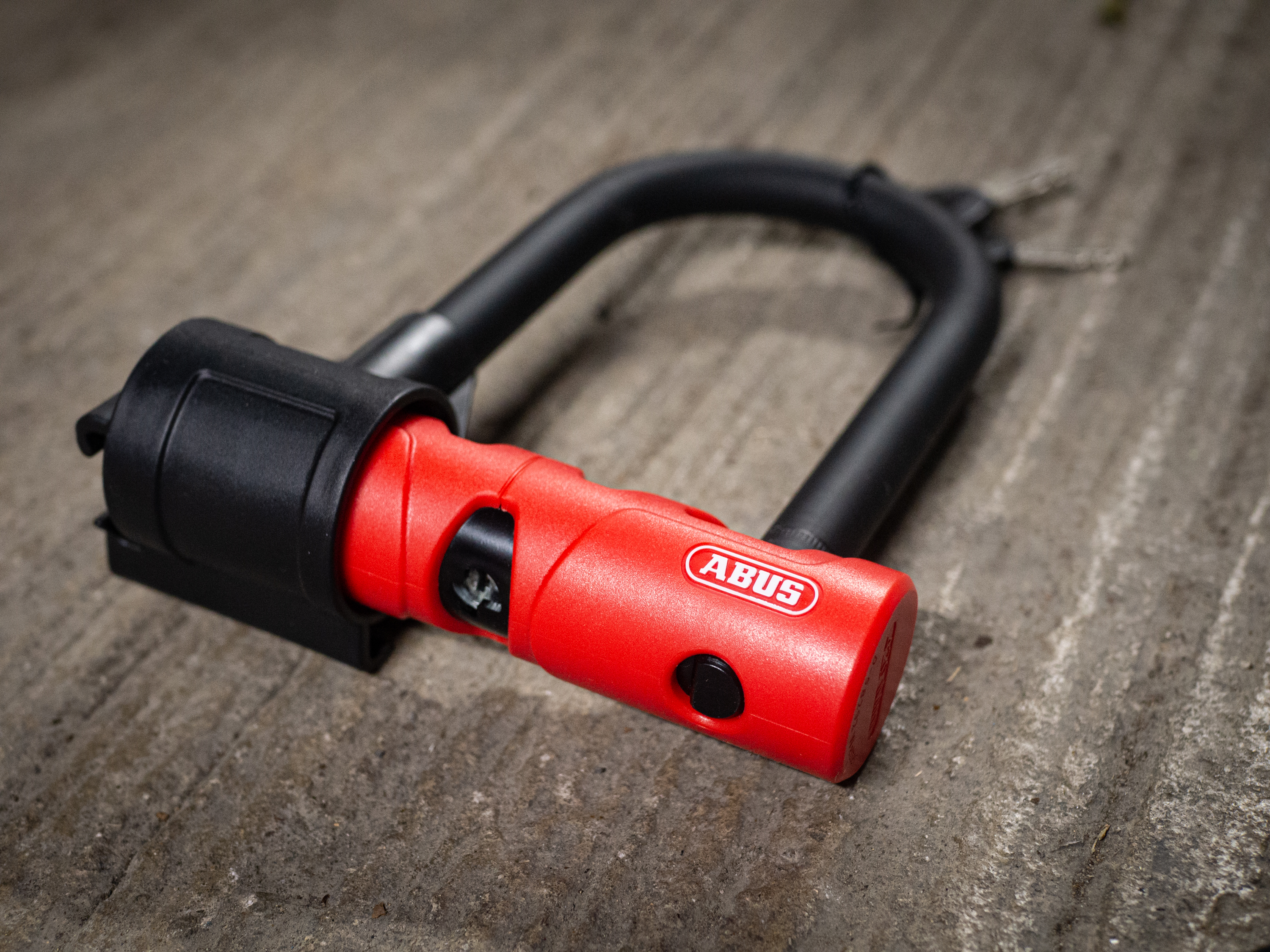 Un candado y un cable Abus Ultimate 420 negro y rojo sobre un suelo de piedra junto a un portabicicletas