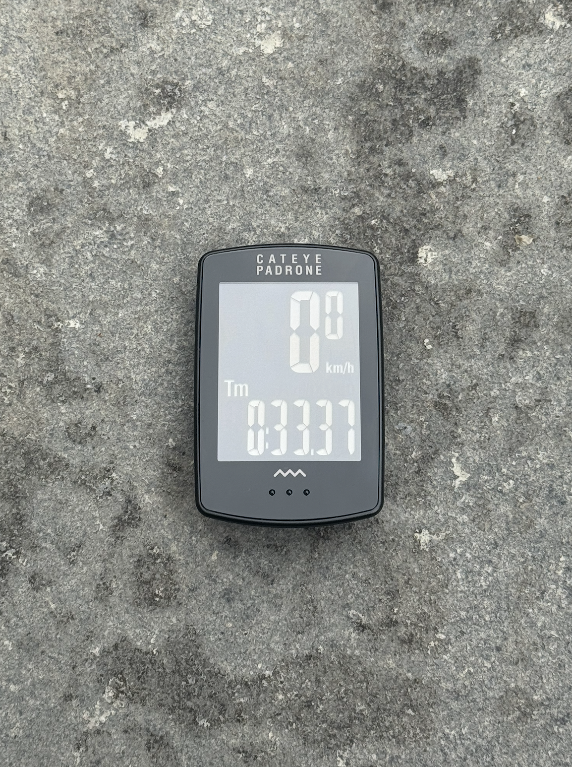 El Padrone Cateye se sienta sobre un fondo gris con la pantalla iluminada y mostrando configuraciones de km/h y tiempo