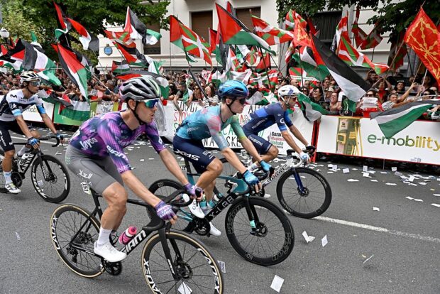 Cyclists compete as pro-Palestinian protesters holding Palestinian and Basque flags demonstrate during the eleventh stage of the Vuelta a Espana cycling tour, a 167 km race from Bilbao to Bilbao, on September 3, 2025. (Photo by ANDER GILLENEA / AFP)