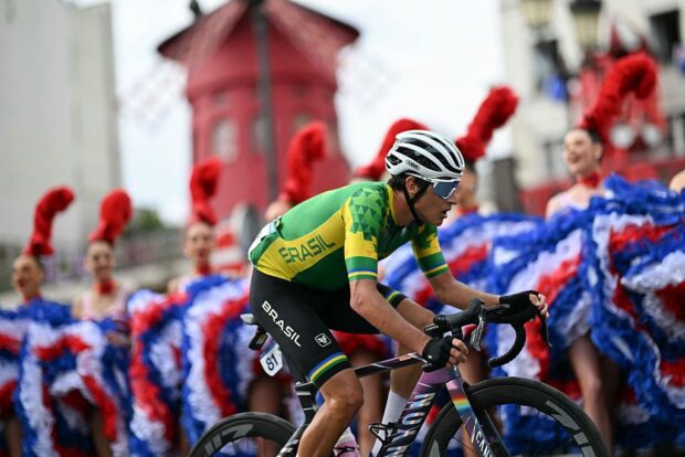 Brazil's Vinicius Rangel Costa cycles past Moulin Rouge dancers performing the cancan outside the Moulin Rouge cabaret during the men's cycling road race during the Paris 2024 Olympic Games in Paris, on August 3, 2024. (Photo by Mauro PIMENTEL / AFP)