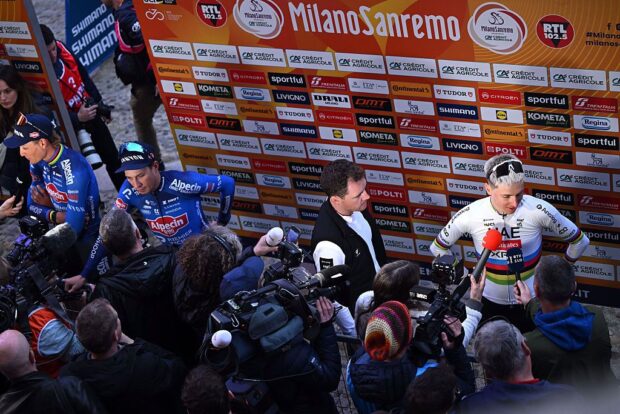 A wide view of the mixed zone with Pogacar in front of the Milan-San Remo sponsor backdrop. Jasper Philipsen and Mathieu van der Poel are being interviewed to his left