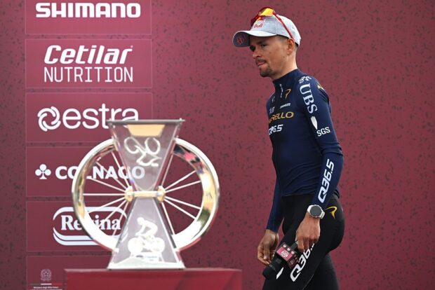Tom Pidcock walks past the Strade Bianche trophy in front of a red backdrop at the teams presentation