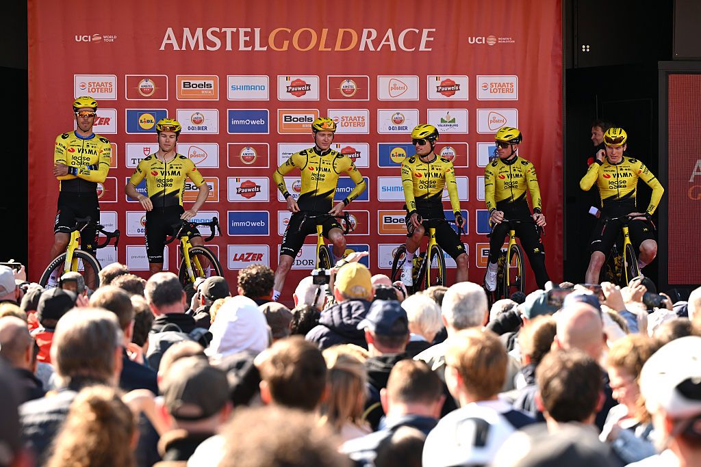 A wide, far away shot of Visma-Lease a Bike riders on the podium for sign on at the start of Amstel Gold Race men's 2026, with the crowd in the foreground