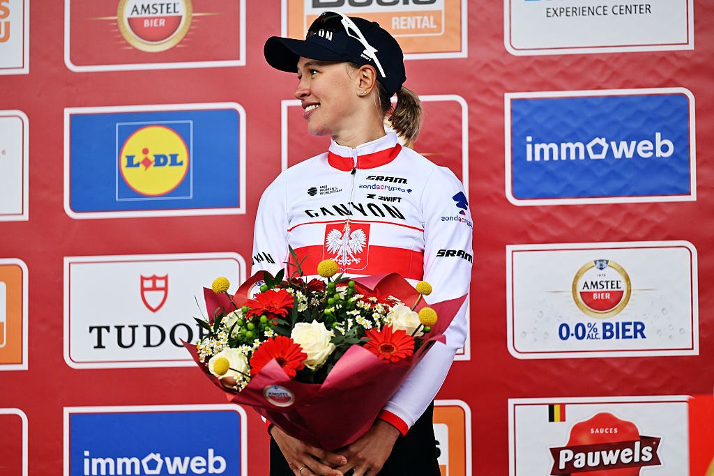 Kasia Niewiadoma-Phinney sube al podio de la Amstel Gold Race 2026, sonriendo a un lado y sosteniendo un ramo de flores frente a ella.