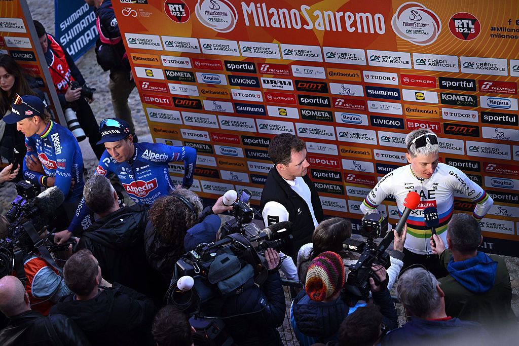 Amplia vista de la zona mixta con Pogacar ante el telón de fondo del patrocinador Milán-San Remo. Jasper Philipsen y Mathieu van der Poel son entrevistados a su izquierda